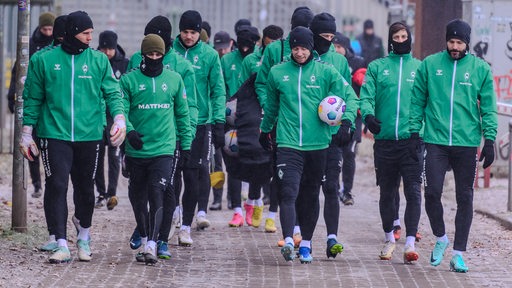 Werder-Spieler sind als Gruppe auf dem Weg zum Trainingsplatz, alle gekleidet mit Trainingsanzügen, schwarzen Wollmützen und Handschuhen.
