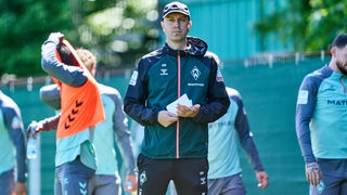 Werder-Coach Ole Werner schaut am Rande des Trainings ernst und hält dabei mehrere Notizzettel in der Hand.
