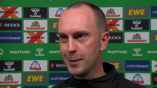 Werder-Trainer Ole Werner steht lächelnd nach dem Spiel gegen Mainz vor einer Werbewand beim Interview.