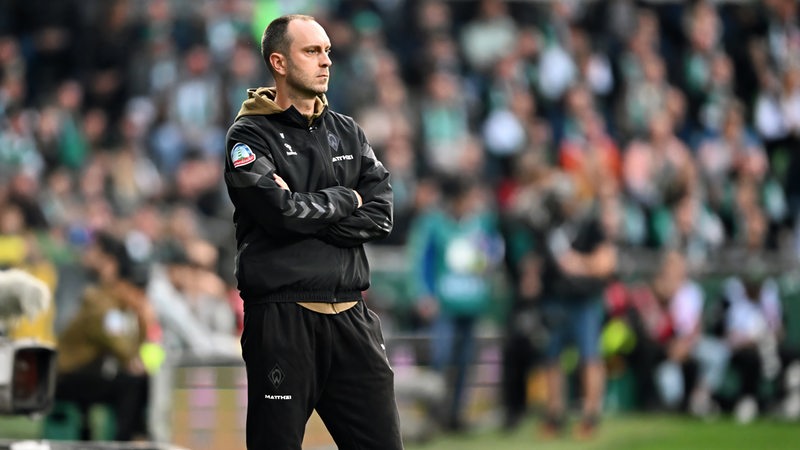 Werder-Trainer Ole Werner steht nachdenklich mit verschränkten Armen am Spielfeldrand.