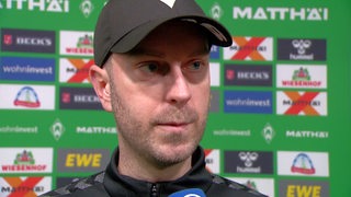 Werder-Trainer Ole Werner trägt eine schwarze Kappe und steht vor einer Werbetafel beim Interview.