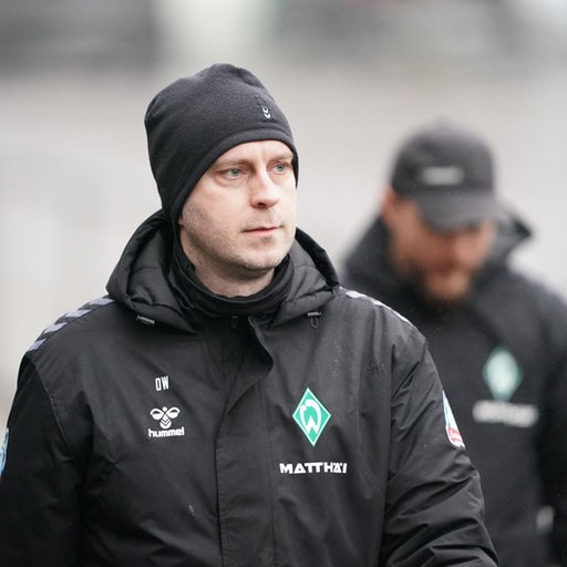 Werder-Trainer Ole Werner mit schwarzer Wollmütze und ernster Miene auf dem Weg zum Trainingsplatz.