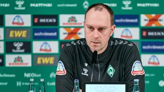 Werder-Trainer Ole Werner sitzt auf dem Podium vor einer grünen Werbewand bei einer Pressekonferenz.