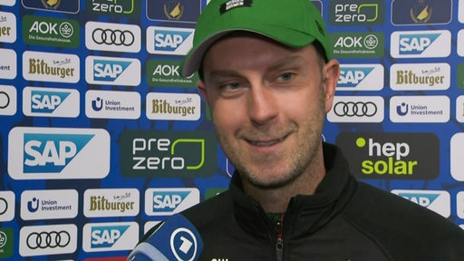 Werder-Trainer Ole Werner steht lächelnd nach dem Spiel gegen Hoffenheim beim Interview vor einer Werbewand.