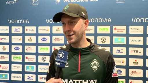 Werder-Trainer Ole Werner steht lächelnd nach dem Spiel in Bochum vor einer Werbewand beim Interview.