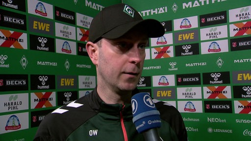 Werder-Trainer Ole Werner steht nach einem Spiel vor einer Werbewand beim Interview und trägt eine schwarze Kappe.