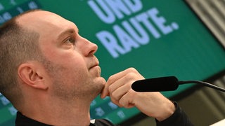 Werder-Trainer Ole Werner im Profil zu sehen während einer Pressekonferenz, er hält sich dabei die Hand ans Kinn.