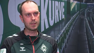 Werder-Trainer Ole Werner sitzt nachdenklich bei einem Interview vor dem Bild der leeren Ostkurve im Weser-Stadion.