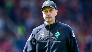 Werder-Trainer Ole Werner geht mit schwarzer Kappe auf dem Kopf konzentriert zu seiner Trainerbank vor einem Spiel.