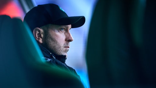 Werder-Trainer Ole Werner sitzt im Halbdunkel auf einer Trainerbank vor einem Spiel.