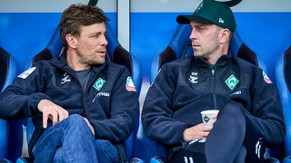Werder-Trainer Ole Werner und Geschäftsführer Clemens Fritz sitzen vor einem Testspiel nebeneinander auf der blauen Trainerbank und schauen sich im Gespräch an.