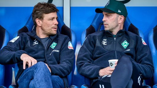 Werder-Trainer Ole Werner und Geschäftsführer Clemens Fritz sitzen vor einem Testspiel nebeneinander auf der blauen Trainerbank und schauen sich im Gespräch an.