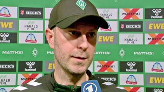 Werder-Trainer Ole Werner steht nach dem Spiel gegen Sankt Pauli beim Interview vor einer Werbewand, seine schwarze Schirmmütze wirft dabei einen dunklen Schatten über seine Augen.