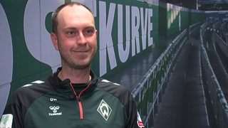 Werder-Trainer Ole Werner sitzt mit einem Lächeln beim Interview vor dem Bild der leeren Ostkurve im Weser-Stadion.