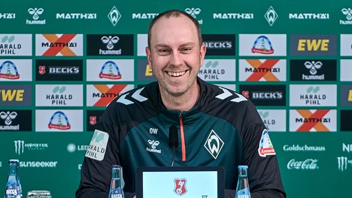 Werder-Trainer Ole Werner sitzt lachend auf dem Podium einer Pressekonferenz vor einer Werbewand.