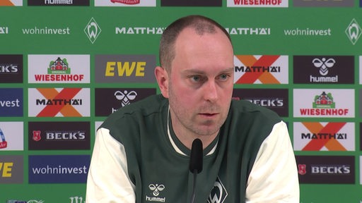 Werder-Trainer Ole Werner sitzt vor einer Werbewand auf dem Podium einer Pressekonferenz.