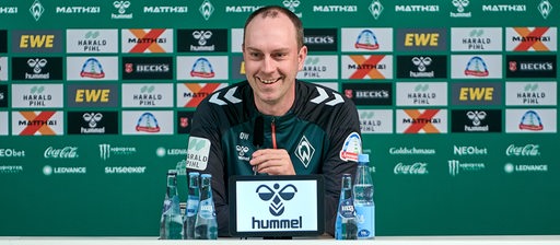 Werder-Trainer Ole Werner lächelt auf dem Podium einer Pressekonferenz, hinter ihm eine Werbewand.