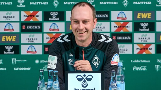 Werder-Trainer Ole Werner lächelt auf dem Podium einer Pressekonferenz, hinter ihm eine Werbewand.