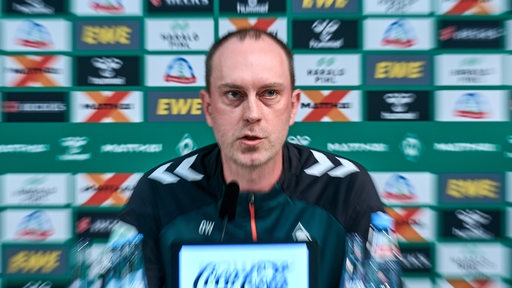 Werder-Trainer Ole Werner sitzt auf dem Podium einer Pressekonferenz vor einer Werbewand, das Bild ist etwas verschwommen als Effekt herangezoomt.