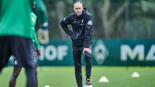 Werder-Coach Ole Werner stützt sich am Rande des Trainings nachdenklich mit dem Fuß auf einen Ball auf.