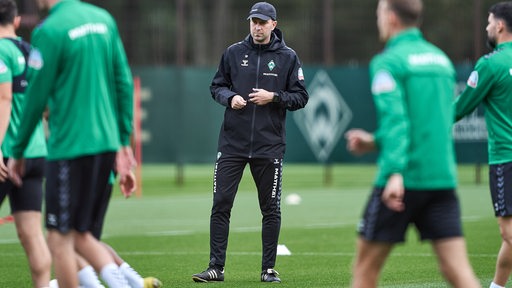 Werder-Trainer Ole Werner staht etwas nachdenklich auf dem Platz, während sich seine Spieler aufwärmen.