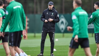 Werder-Coach Ole Werner steht nachdenklich am Rande des Trainings während sich seine Spieler warmlaufen.