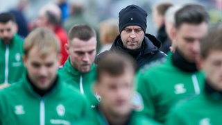 Werder-Coach Ole Werner geht mit leicht gesenktem Kopf und einer schwarzen Mütze gemeinsam mit seinen Spielern zum Trainingsplatz.