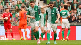 Werder-Spieler Anthony Jung mit hängendem Kopf auf dem Spielfeld, Mitspieler Milos Veljkovic tätschelt ihm im Vorbeigehen den Kopf.