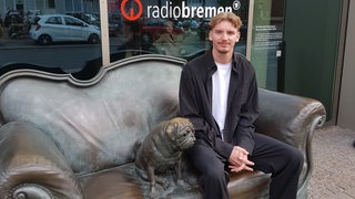 Werder-Profi Nick Woltemade sitzt auf dem Loriot-Sofa vor Radio Bremen neben dem Mops.