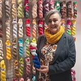 Eine Frau hält Garn in der Hand und steht vor einer Wand, an der Wollknäuel hängen.
