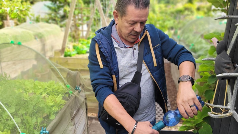 Ein Mann steht in einem Garten und hält eine Plastikflasche in der Hand.