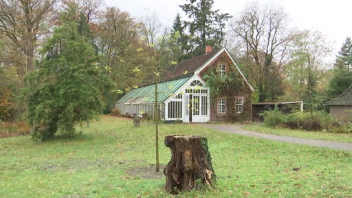 Ein altes Gebäude mit Gewächshaus steht in einem Garten zwischen einigen Bäumen.