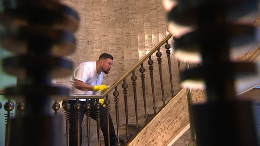 Ein Mann putzt mit gelben Handschuhen ein Treppengeländer einer Wendeltreppe. 