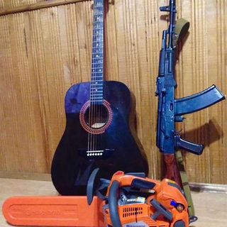 Eine Gitarre, ein Kalashinkov-Gewehr lehnen an einer Holzwand. Am Boden liegt eine Motorsäge. 