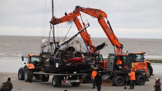 Zwei Bagger heben ein Schiffswrack am Strand auf einen Tieflader.