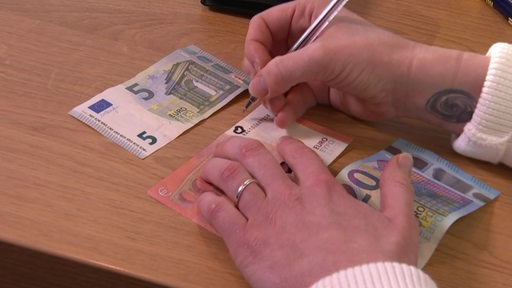 Auf einem Tisch liegen einige Geldscheine, eine Person schreibt etwas darauf.