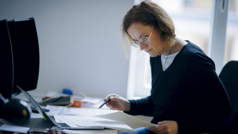 Eine Frau sitzt am Schreibtisch im Buero und liest in Unterlagen. 