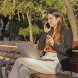 Eine junge Geschäftsfrau sitzt mit ihrem Laptop telefonierend auf einer Parkbank.