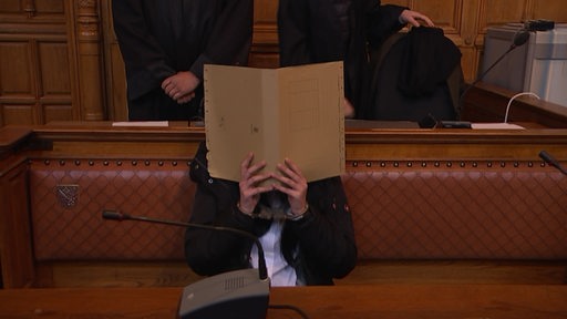 Eine Frau in einem Gerichtssaal. Sie verdeckt ihr Gesicht mit einer Pappmappe.