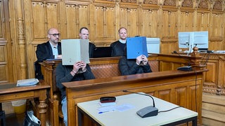 Zwei Angeklagte halten sich Mappen vor ihre Gesichter. Im Hintergrund sitzen drei Anwälte und schauen in den Gerichtssaal.