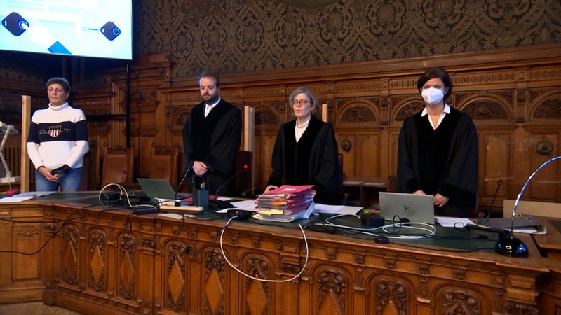 In einem Gerichtssaal stehen zwei Männer und zwei Frauen. Vor ihnen liegen Aktenordner und Laptops.
