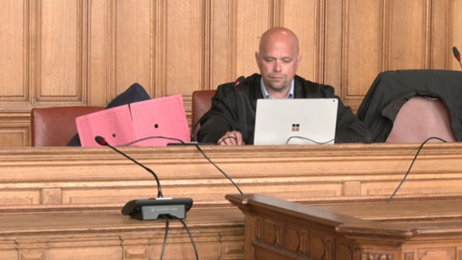 Eine Person sitzt in einem Gericht und verdeckt das Gesicht mit einer Mappe. Neben ihr sitzt ein Richter vor einem Laptop.