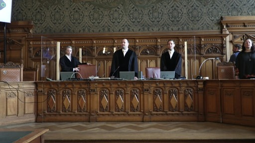 Drei Richter stehen in einem Gerichtssaal an ihren Plätzen.