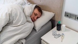 Ein Mann liegt krank im Bett. auf dem Nachttisch liegen ein Fieberthermometer und Tabletten.