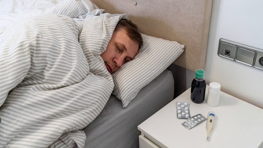 Ein Mann liegt krank im Bett. auf dem Nachttisch liegen ein Fieberthermometer und Tabletten.