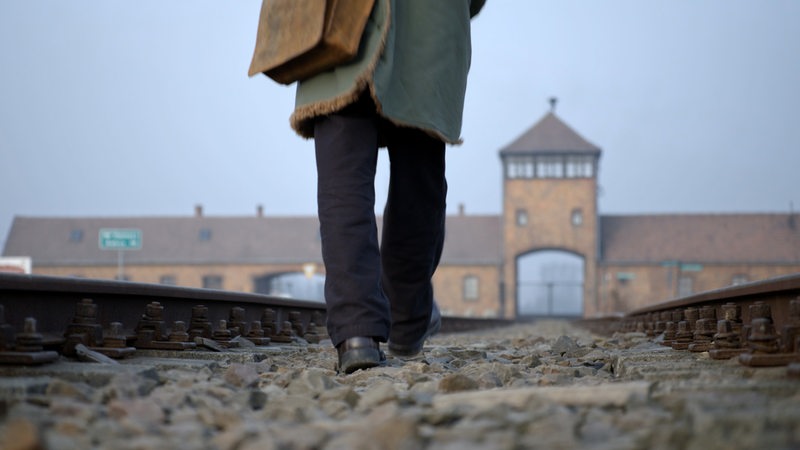 Eine Person geht auf den Gleisen Richtung Zugeinfahrtstor des ehemaligen KZ Auschwitz-Birkenau (Filmszene)