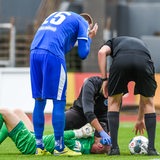 Ein Torwart liegt nach einem Foulspiel verletzt am Boden. Schiedsrichter, Sanitäter und ein Spieler stehen daneben.