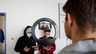 Zwei junge Menschen mit einem Mundschutz halten ein Tablet in der Hand und werden dabei gefilmt