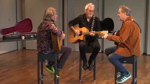 Die Drei Rocklegenden der Band "The Hoolies" sitzen sich mit ihren Gitarren gegenüber.
