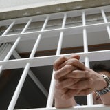 Aus einem vergitterten Fenster streckt ein Mann seine gefalteten Hände heraus.